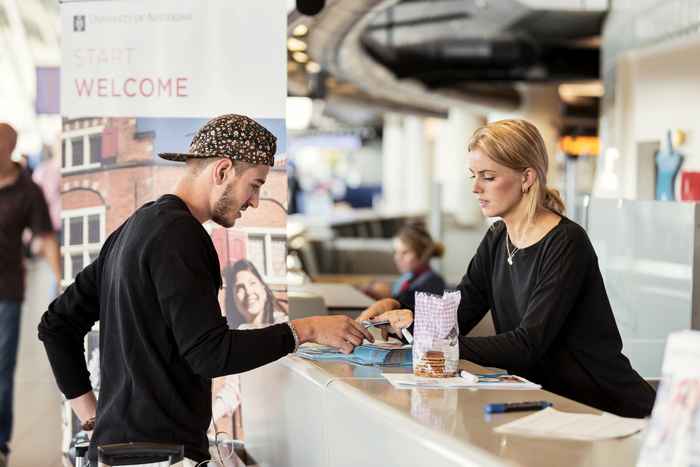 An international student who has just arrived at Schiphol Airport stands at a desk and is welcomed by a host of the University of Amsterdam