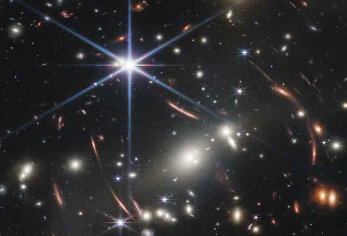 Een foto vol met sterrenstelsels tegen een donkere achtergrond. De sterrenstelsels zijn wollige neveltjes, het licht van somme ervan zijn uitgerekt. Dit komt door de zwaartekracht van de sterenstelsels op de voorgrond.