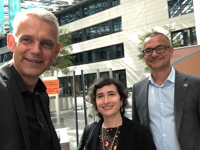 Photo of a smiling Heino Faclke, Sera Markoff and Rob Fender. Heino Falcke is a tall white man with gray hair and is wearing a dark jacket. Sera Markoff is a petite woman with medium length curls and a black hole dress. Rob Fender is slightly shorter than Heino, wears glasses and his hair is cropped, and he wears a gray jacket.