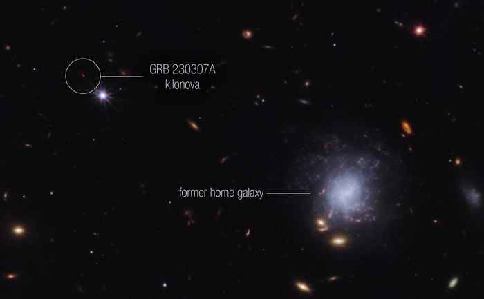 Een zwarte achtergrond met rechts onderin een cluster sterrenstelsels, vlekjes in het blauw en geel. Het grootste sterrenstelsel is blauwpaars heeft de vorm van een draaikolk. Linksbovenin staat een relatief heldere rode stip. Dit is de gammaflits die uit het grote sterrenstelsel rechtsonderin komt.