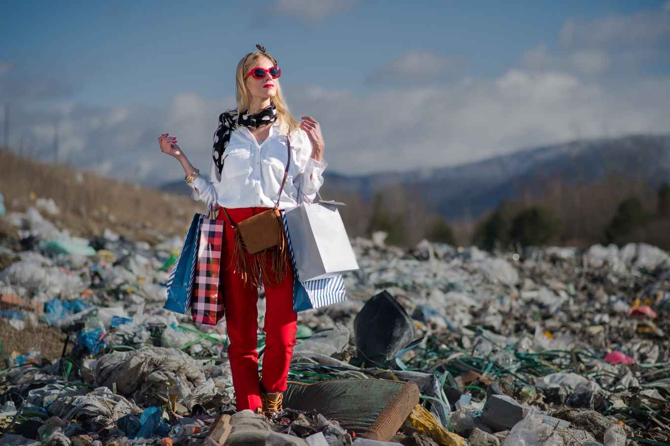 vrouw staat op een grote afvalberg terwijl ze haar 'shopping bags' vasthoudt